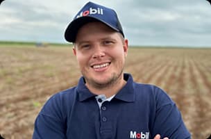 João Pierobon usando camisa polo azul e boné em área rural com campo ao fundo.