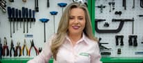 Sandra Nalli, fundadora da Escola do Mecânico, usando camisa branca em ambiente de oficina com equipamentos verdes ao fundo.