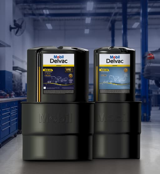 Tambores e galões Mobil Delvac em oficina mecânica com veículos ao fundo.