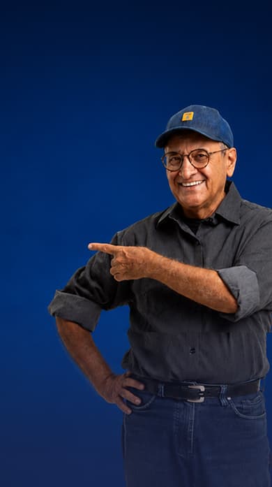 Homem sorrindo usando boné azul, óculos e camisa cinza, apontando para o lado sobre fundo azul.