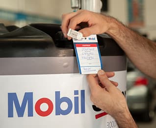 Mãos aplicando etiqueta de identificação em tanque de óleo Mobil dentro de oficina automotiva.